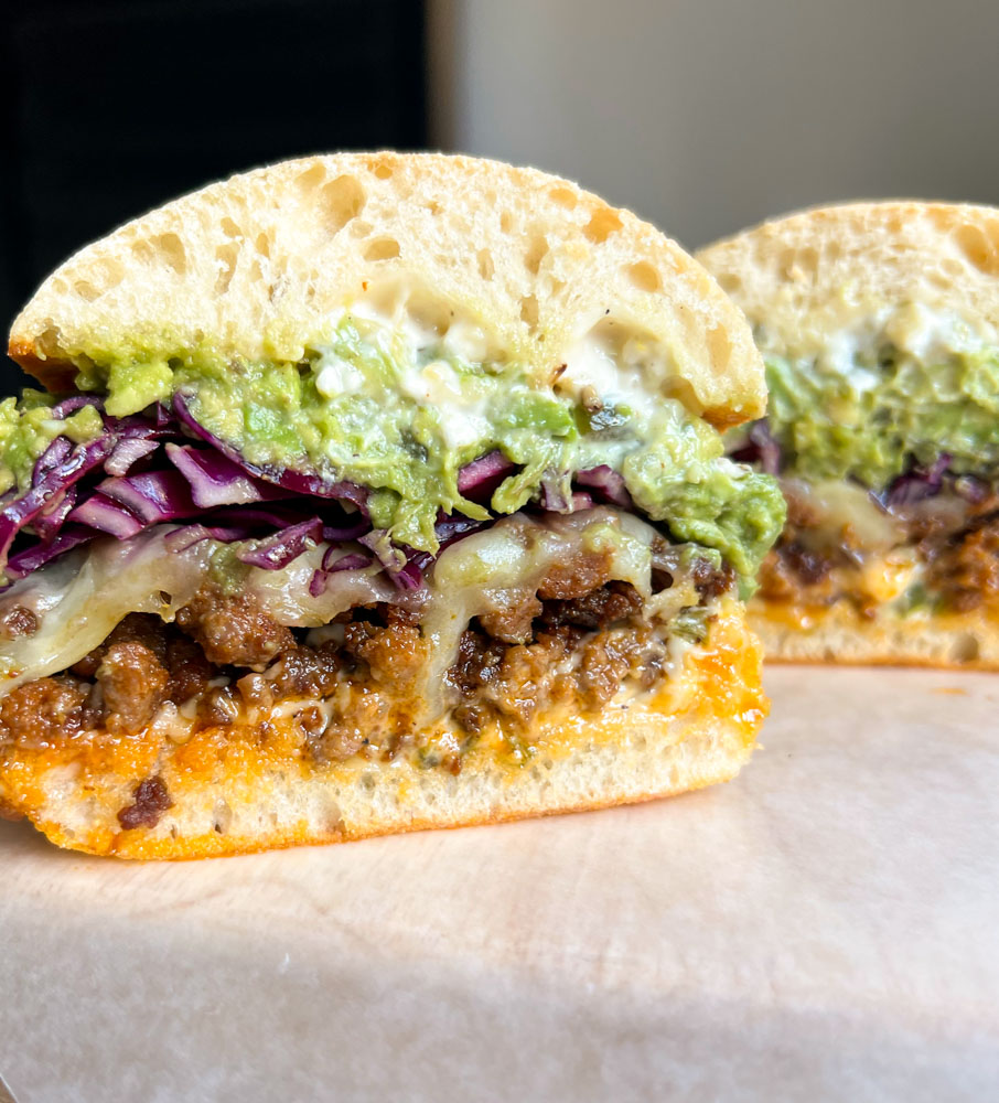 close up of chorizo sandwich cut in half with melted cheese, red cabbage, mashed avocado 