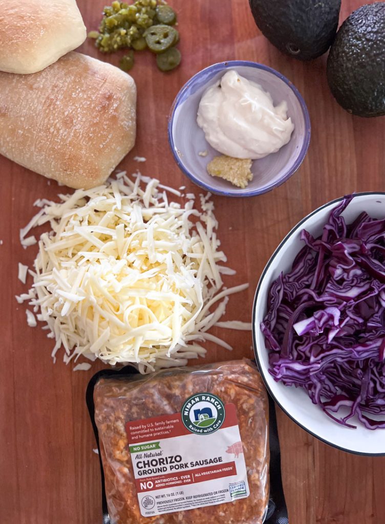 cutting board with shredded cheese, bread rolls, pickled jalape&ntilde;o slices, two whole avocados, small bowl with sour cream and minced garlic, bowl with chopped red cabbage, one pack of uncooked ground chorizo.