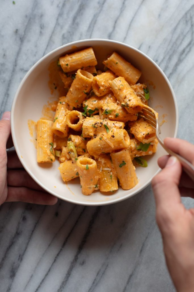 hand holding bowl of rigatoni pasta and other hand using a fork