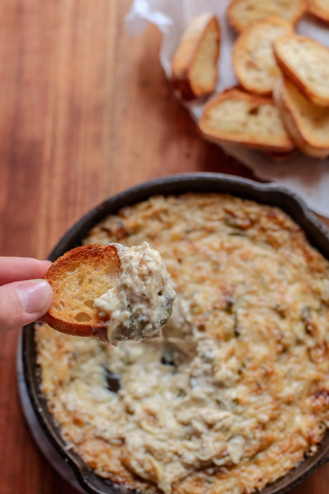 hand holding crostini that's been dipped in artichoke dip with cast iron skillet filled with dip in background and bowl of crostini
