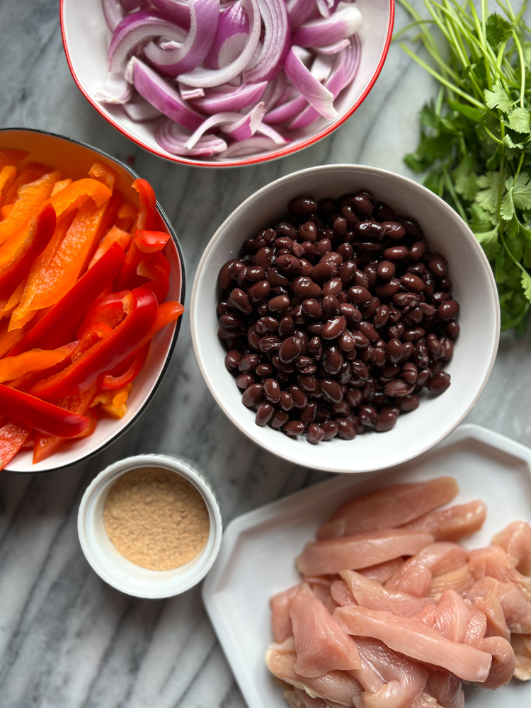 prepped ingredients for chicken fajita wraps: sliced peppers, sliced red onion, bowl of black beans, sliced chicken breast, cilantro, fajita seasoning.