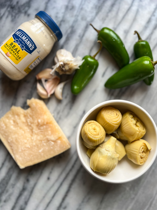 ingredients on counter to make artichoke dip: jar of mayo, garlic cloves, jalape&ntilde;os, parmesan cheese, and artichokes in a bowl.
