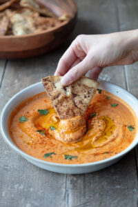 shallow bowl with hand dipping pita chip with bowl of pita chips in background