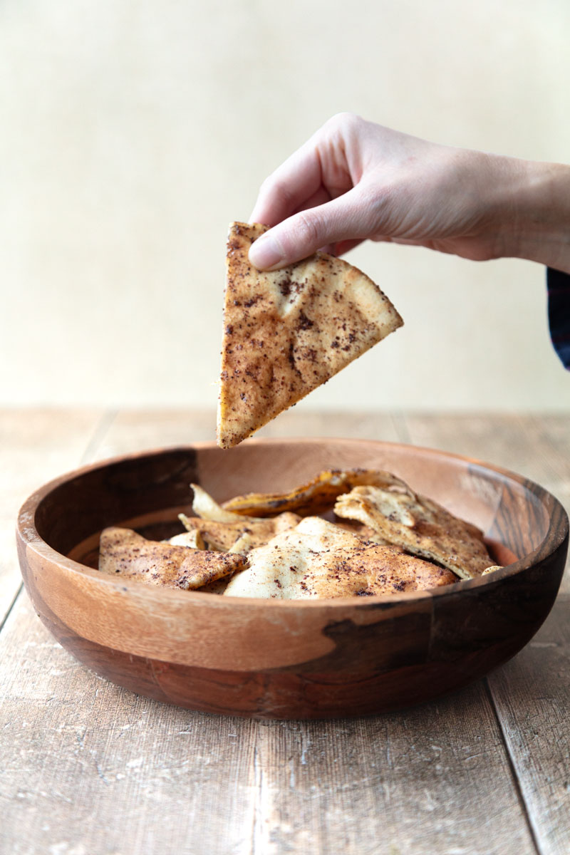 hand holding pita chip over wooden bowl filled with pita chips