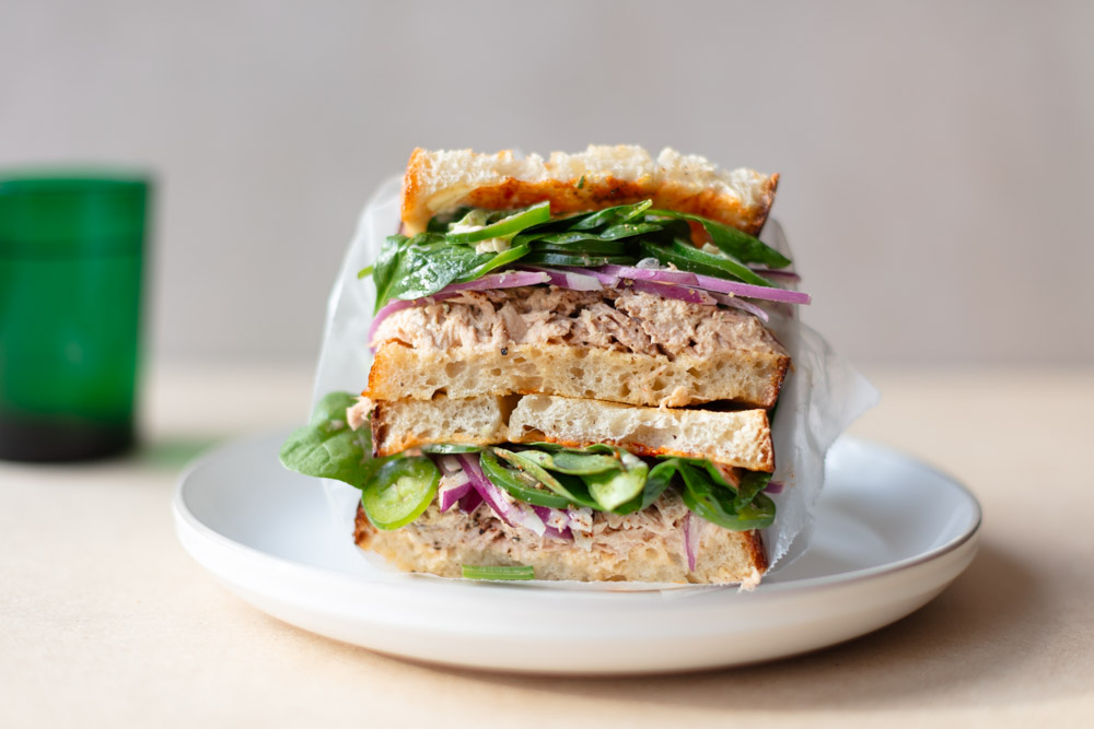 Stacked spicy tuna sandwich with Sriracha mayo, baby spinach, and red onion on toasted whole grain bread.