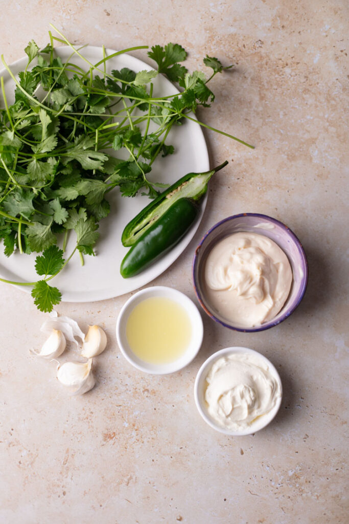 fresh cilantro and jalapeno pepper on a plate, garlic cloves, lime juice in small bowl, sour cream, and mayo