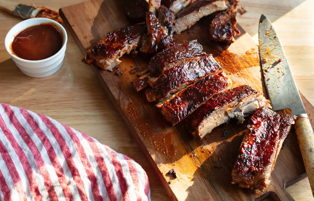 bbq ribs cut in pieces on a cutting board with a knife and small bowl of sauce on the side