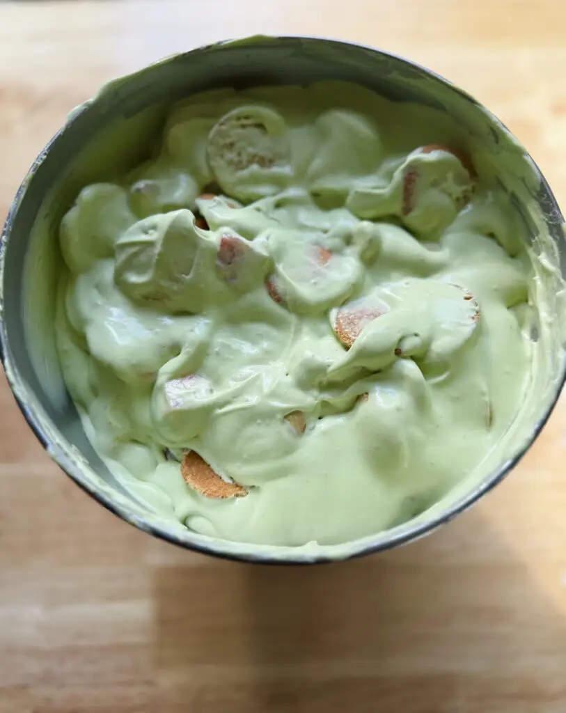 Nilla wafers, strawberries and bananas folded into the matcha whipped cream mixture in a mixing bowl.