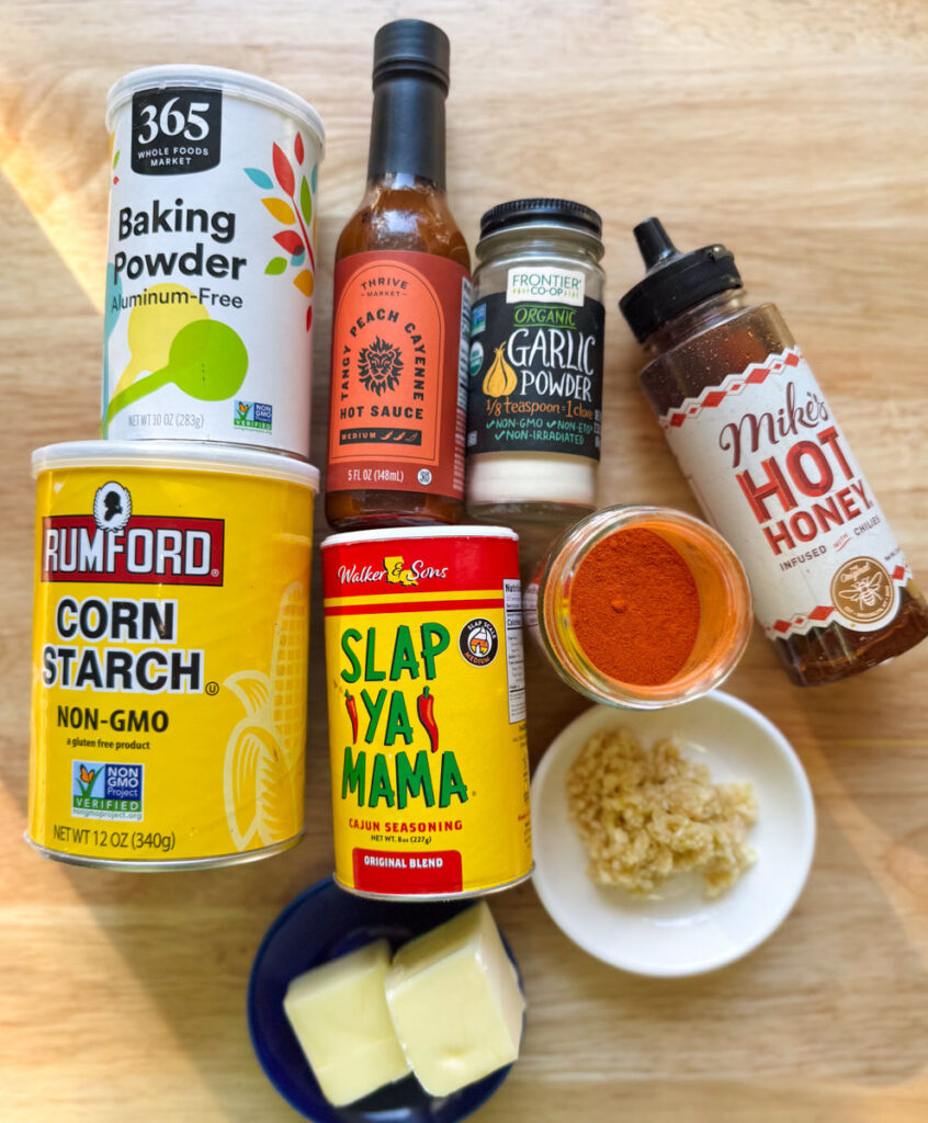 Ingredients for hot honey garlic chicken wings, including cornstarch, baking powder, hot honey, garlic, butter, and Cajun seasoning, on a wooden surface.