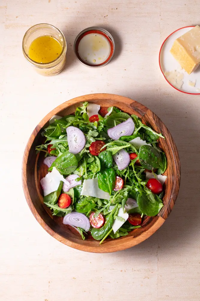 Overhead shot of spinach arugula salad next to a jar of dressing and a block of parmesan cheese.