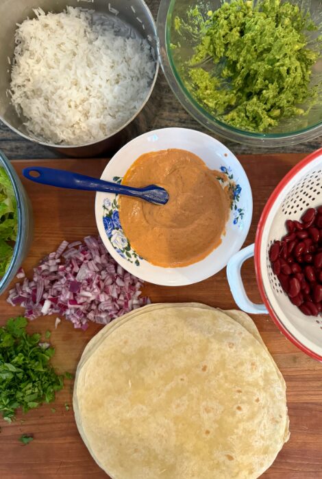 Flatlay of fresh burrito ingredients including flour tortillas, cooked white rice, guacamole, seasoned creamy sauce, red kidney beans, chopped red onions, fresh cilantro, and shredded lettuce&mdash;perfect for assembling cheesy beef and bean burritos at home.