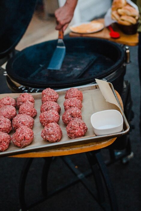 Raw ground beef burger patties lined up on a baking tray beside a grill, with a person holding a spatula and burger buns in the background, ready for a backyard cookout.
