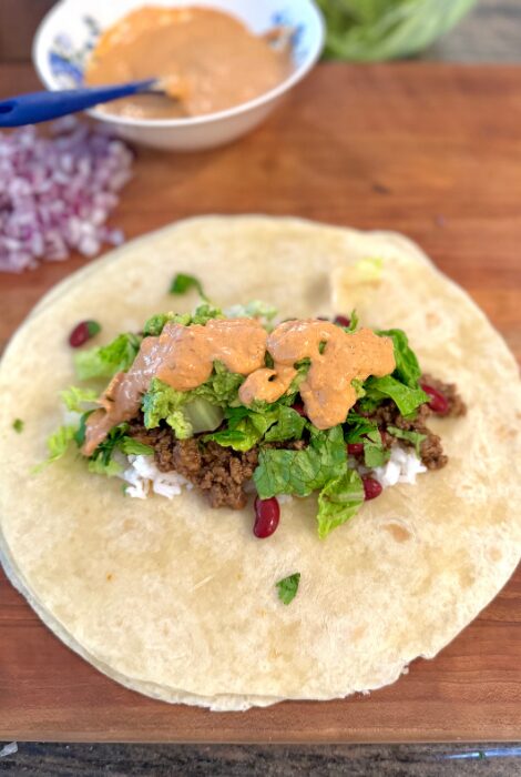 Open Cheesy Beef & Bean Burrito on a wooden board, layered with seasoned ground beef, kidney beans, rice, chopped lettuce, red onion, guacamole, and a drizzle of creamy chipotle sauce, ready to be wrapped.