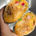 A hand holds two halves of a cheesy beef and bean burrito wrapped in foil, revealing a delicious cross-section filled with seasoned ground beef, melty cheese, rice, beans, and fresh vegetables inside a warm tortilla.