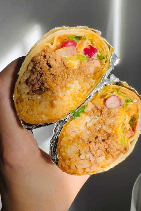 Close-up of a hand holding two foil-wrapped cheesy beef and bean burrito halves, showing a savory filling of seasoned ground beef, melted cheese, rice, beans, lettuce, and chopped veggies, perfect for meal prep, freezer storage, or teen-friendly dinners.