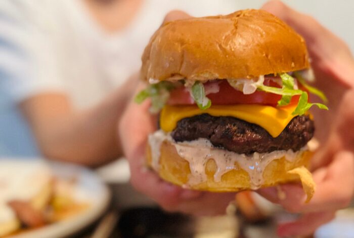 A juicy cheeseburger with awesome sauce topped with melty cheddar, shredded lettuce, tomato, and creamy sauce, held over a tray in a casual home setting.