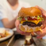 Close-up of a hand holding a juicy cheeseburger with melted cheddar, lettuce, tomato, and creamy sauce, with fries on a tray in the foreground and a smiling teen in the background.