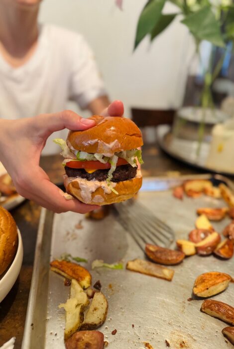 Hand holding a juicy cheeseburger layered with cheddar, tomato, pickles, shredded lettuce, and awesome sauce on a brioche bun, served over a tray with crispy roasted potatoes in a casual kitchen setting.