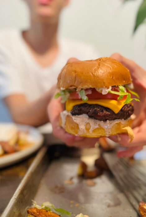 Close-up of a hand holding a juicy cheeseburger with melted cheddar, lettuce, tomato, and creamy sauce, with fries on a tray in the foreground and a smiling teen in the background.
