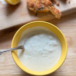 Homemade blue cheese dressing in a yellow bowl with visible crumbles and a spoon, served beside crispy breaded chicken and lemon wedges.