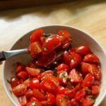 a close-up of a spoonful of marinated cherry tomatoes lifted from a bowl, showing the glistening mix of olive oil, herbs, and balsamic, with fresh baguette slices blurred in the background