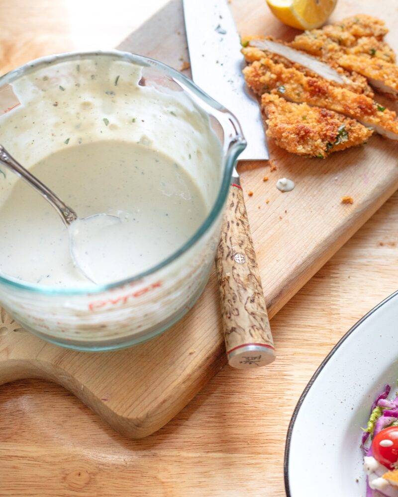 blue cheese dressing in Pyrex with spoon, crispy chicken slices, lemon wedge, and knife on wooden cutting board