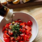balsamic vinegar being poured over a bowl of halved cherry tomatoes mixed with minced garlic, chopped herbs, cracked pepper, and olive oil, with fresh baguette slices in the background