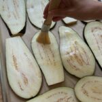 hand brushing sliced eggplant with olive oil on a parchment-lined tray