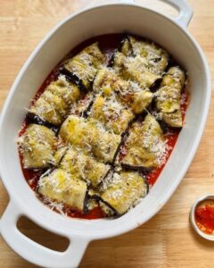 rolled eggplant slices nestled in tomato sauce inside a white baking dish, topped with grated cheese and herbs, ready for the oven