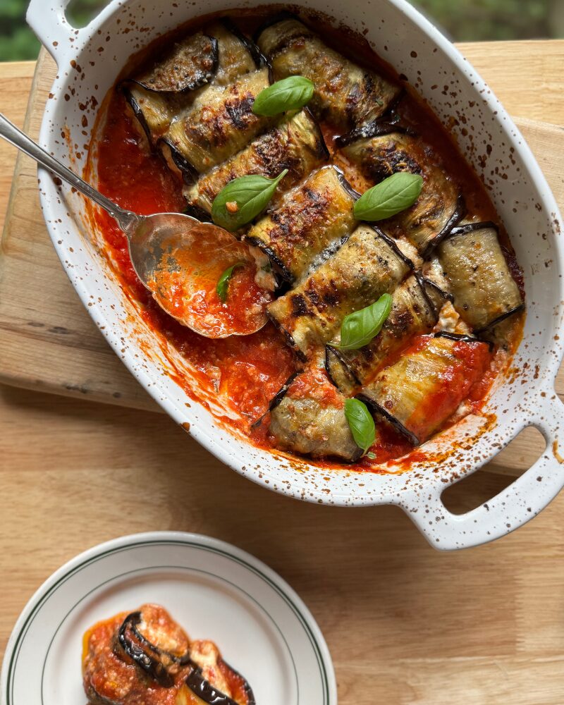 a serving of eggplant rollatini plated beside a baking dish filled with more rollatini in rich tomato sauce, garnished with fresh basil