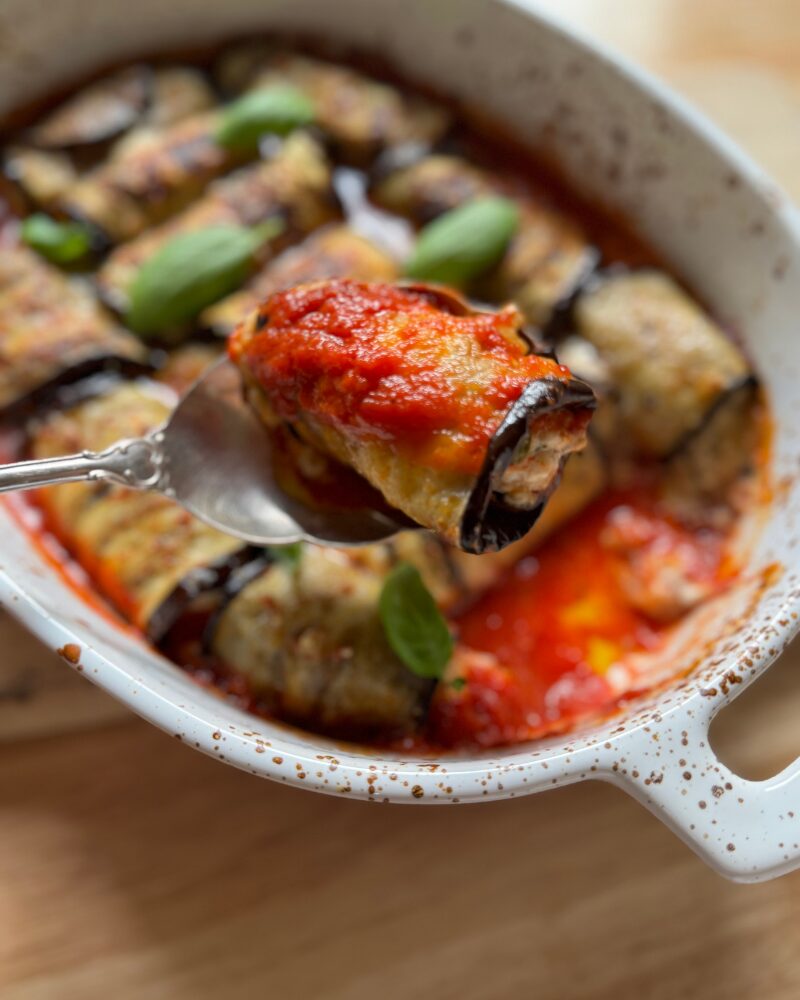 a close-up of a spoon lifting a saucy eggplant rollatini from a white baking dish, revealing its cheesy filling and vibrant tomato sauce