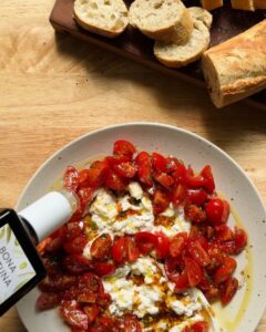 a bottle of olive oil being drizzled over a vibrant tomato and burrata dip, surrounded by marinated cherry tomatoes and finished with vinaigrette, with sliced baguette in the background