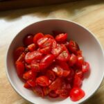 a white bowl filled with freshly halved cherry tomatoes on a wooden table, with slices of crusty baguette visible in the background