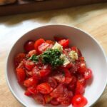 a bowl of halved cherry tomatoes topped with minced garlic, chopped parsley, cracked black pepper, and seasonings, ready to be mixed for a fresh tomato marinade. Slices of baguette rest in the background on a wooden cutting board