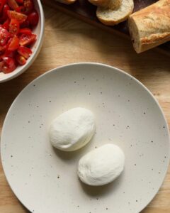 two whole burrata balls placed on a speckled ceramic plate, with a bowl of marinated cherry tomatoes and sliced baguette visible in the background, ready for assembly
