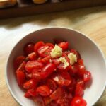 a bowl of halved cherry tomatoes topped with freshly minced garlic, set on a wooden table with sliced baguette pieces in the background