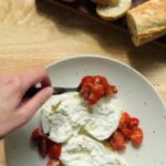 a hand spooning marinated cherry tomatoes over torn burrata on a speckled ceramic plate, with fresh baguette slices visible in the background
