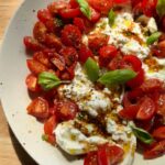 a fresh tomato and burrata dip served on a plate, topped with marinated cherry tomatoes, a drizzle of vinaigrette, cracked black pepper, and garnished with whole basil leaves
