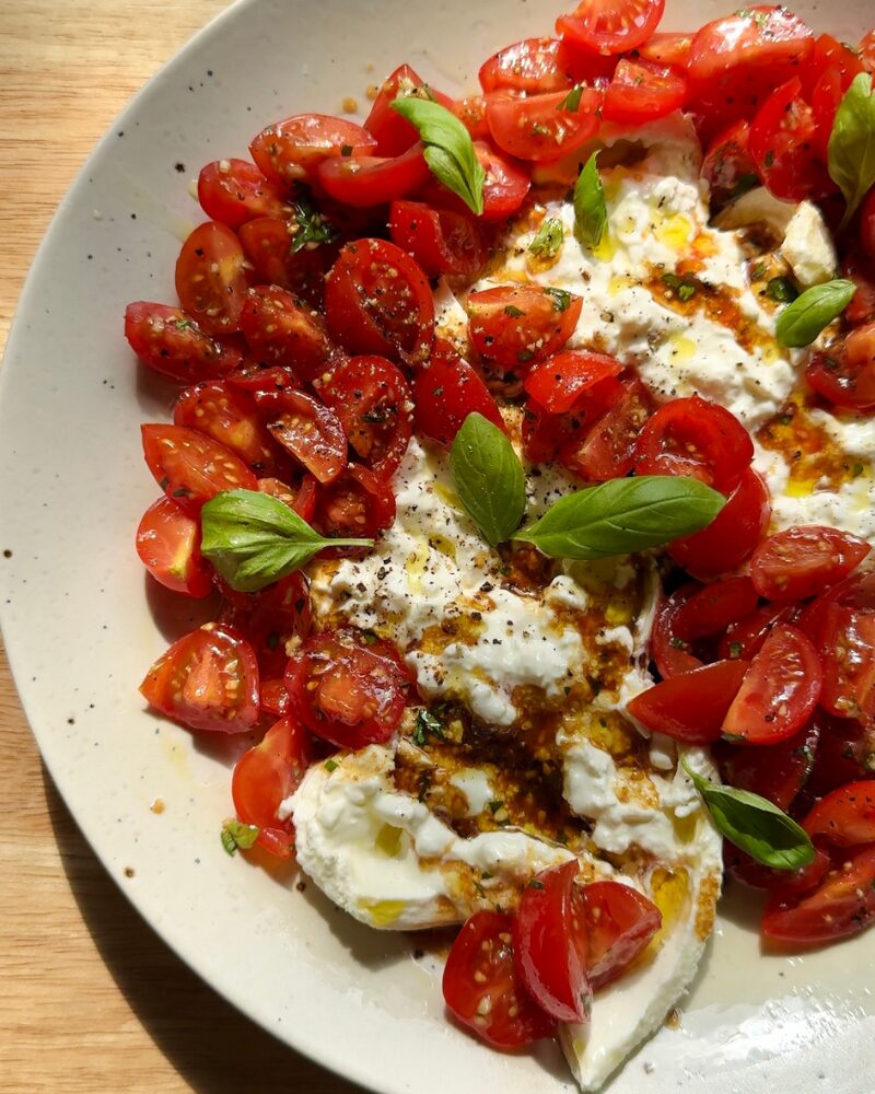 a fresh tomato and burrata dip served on a plate, topped with marinated cherry tomatoes, a drizzle of vinaigrette, cracked black pepper, and garnished with whole basil leaves