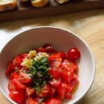 a bowl filled with halved cherry tomatoes, fresh minced garlic, chopped herbs, cracked black pepper, and a drizzle of olive oil, ready to be mixed for a tomato topping. Sliced baguette sits on a wooden board in the background