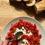 a hand holding a spoon drizzling tomato vinaigrette over a plate of creamy burrata and marinated cherry tomatoes, with sliced baguette in the background