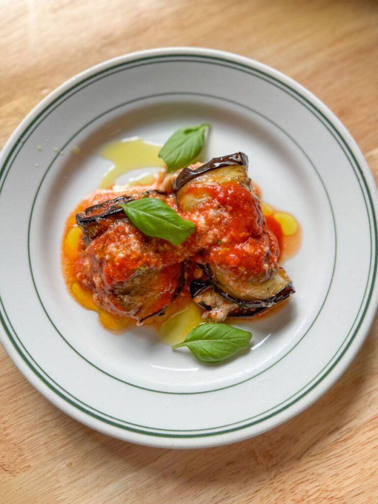 Rolled eggplant slices layered with tomato sauce and melted cheese, topped with fresh basil leaves and a drizzle of olive oil on a white plate.