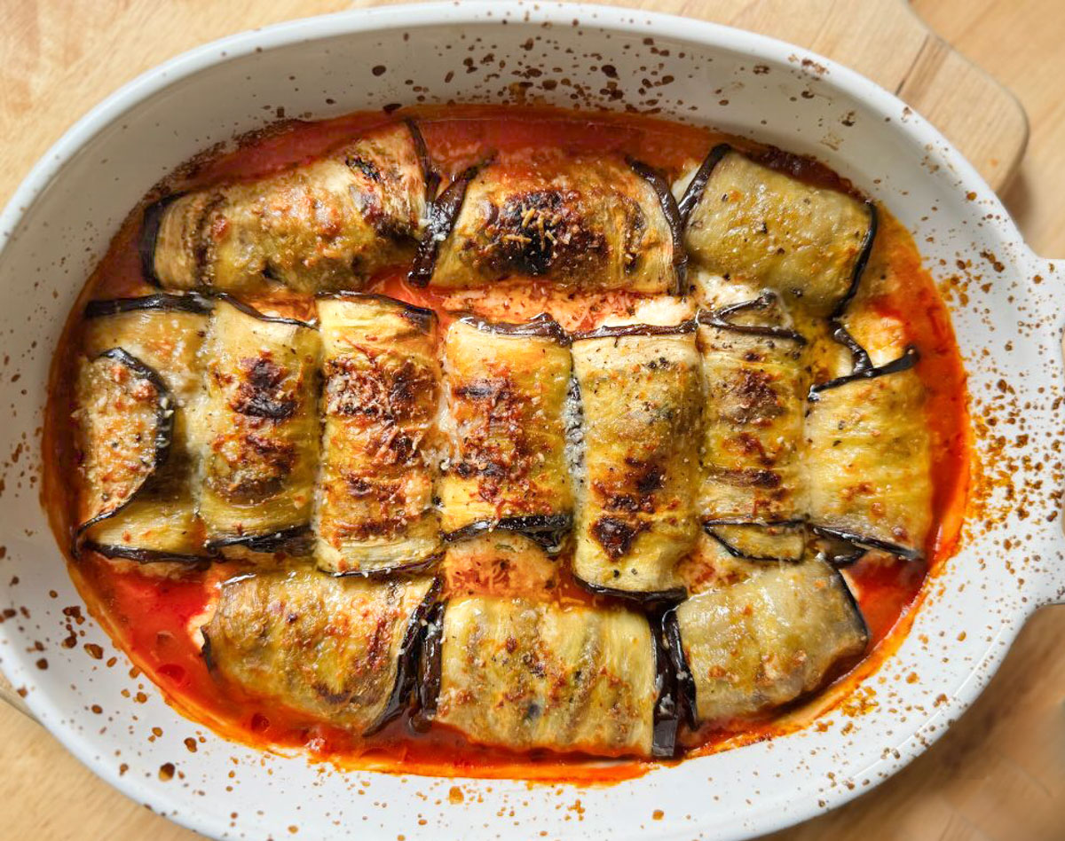 Baked eggplant involtini arranged in a white oval dish, filled and rolled, topped with tomato sauce, melted cheese, and a sprinkle of herbs.