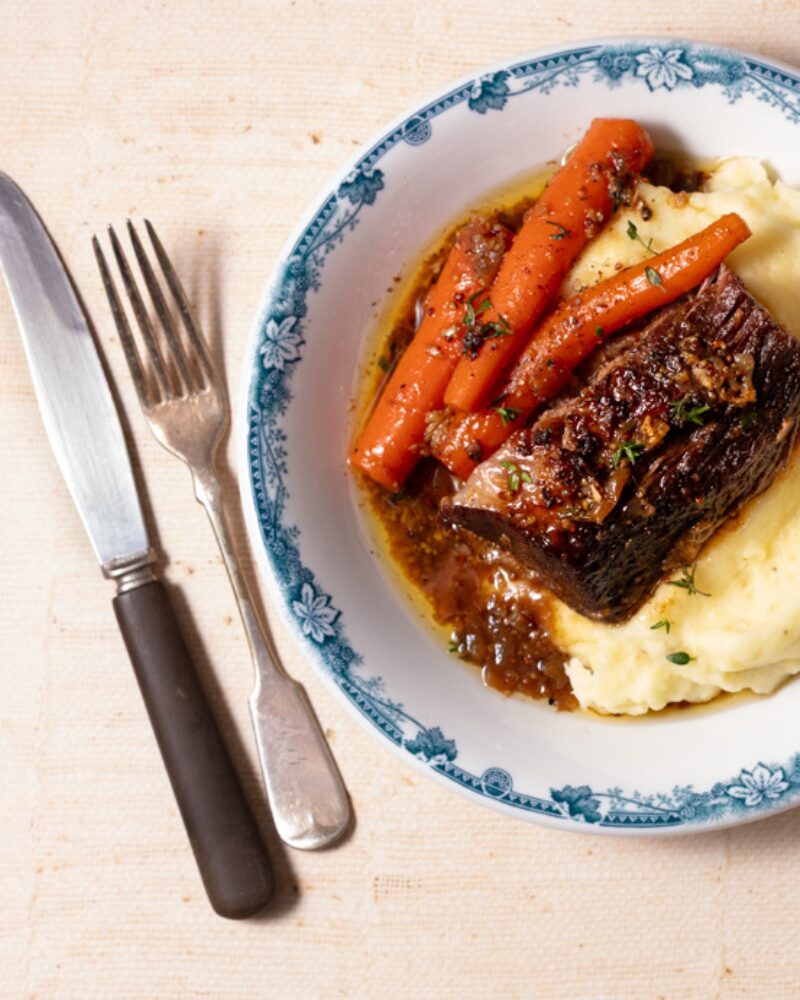 plated braised beef with tender carrots and creamy mashed potatoes in a rich pan sauce, set on a blue-rimmed plate with fork and knife alongside