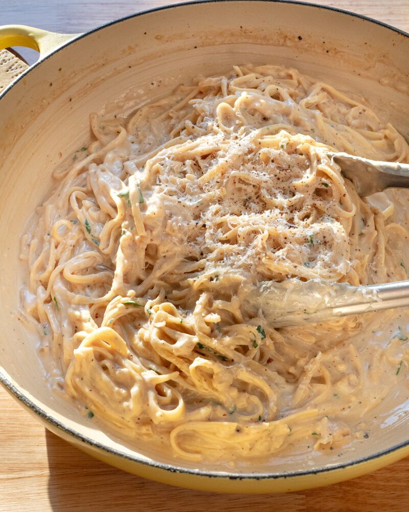 creamy parmesan pasta being gently tossed in a skillet with a rich, velvety cheese sauce, freshly grated parmesan, cracked black pepper, and flecks of fresh herbs