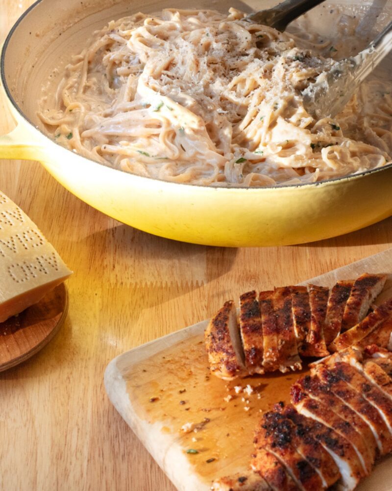 creamy parmesan pasta tossed in a silky cheese sauce inside a yellow skillet, with freshly grated parmesan and herbs, alongside sliced seared chicken breast resting on a wooden board
