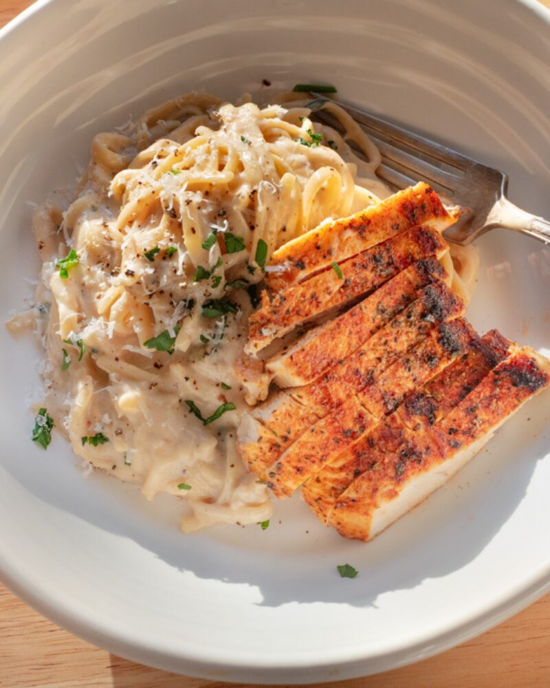 creamy parmesan pasta twirled in a rich, silky cheese sauce with freshly cracked black pepper and herbs, served in a white bowl alongside sliced seared chicken breast