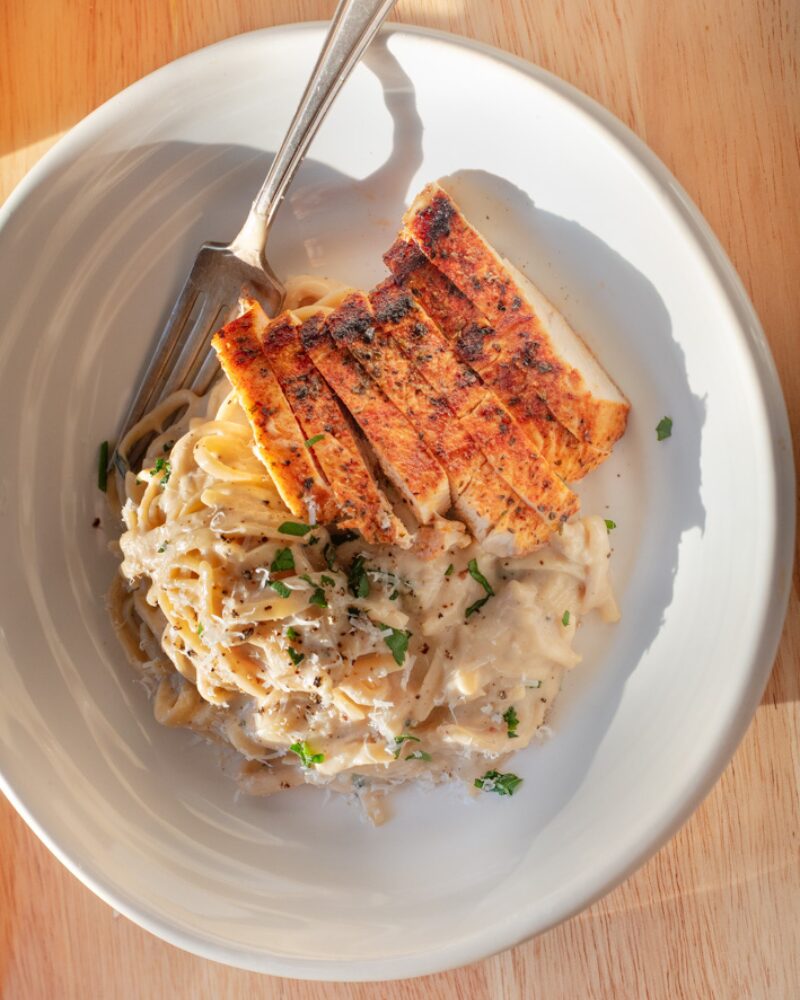 creamy parmesan pasta tossed in a rich, velvety cheese sauce with cracked black pepper and fresh herbs, topped with sliced seared chicken breast in a white bowl on a wooden table