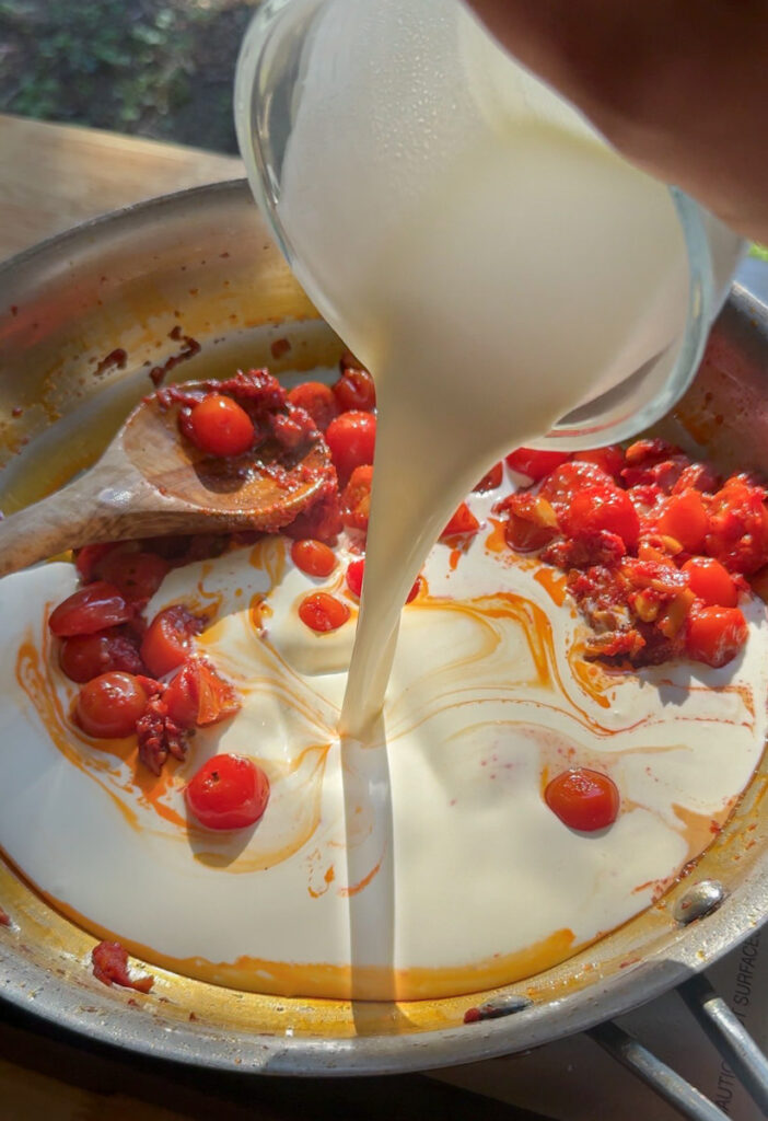 heavy cream being poured into tomato paste and cherry tomatoes in skillet for creamy lobster ravioli sauce