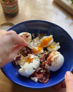 soft boiled eggs with jammy yolk breaking open, topped with crispy bacon, chili crisp, mayonnaise, and fresh herbs in blue bowl for spicy egg salad recipe